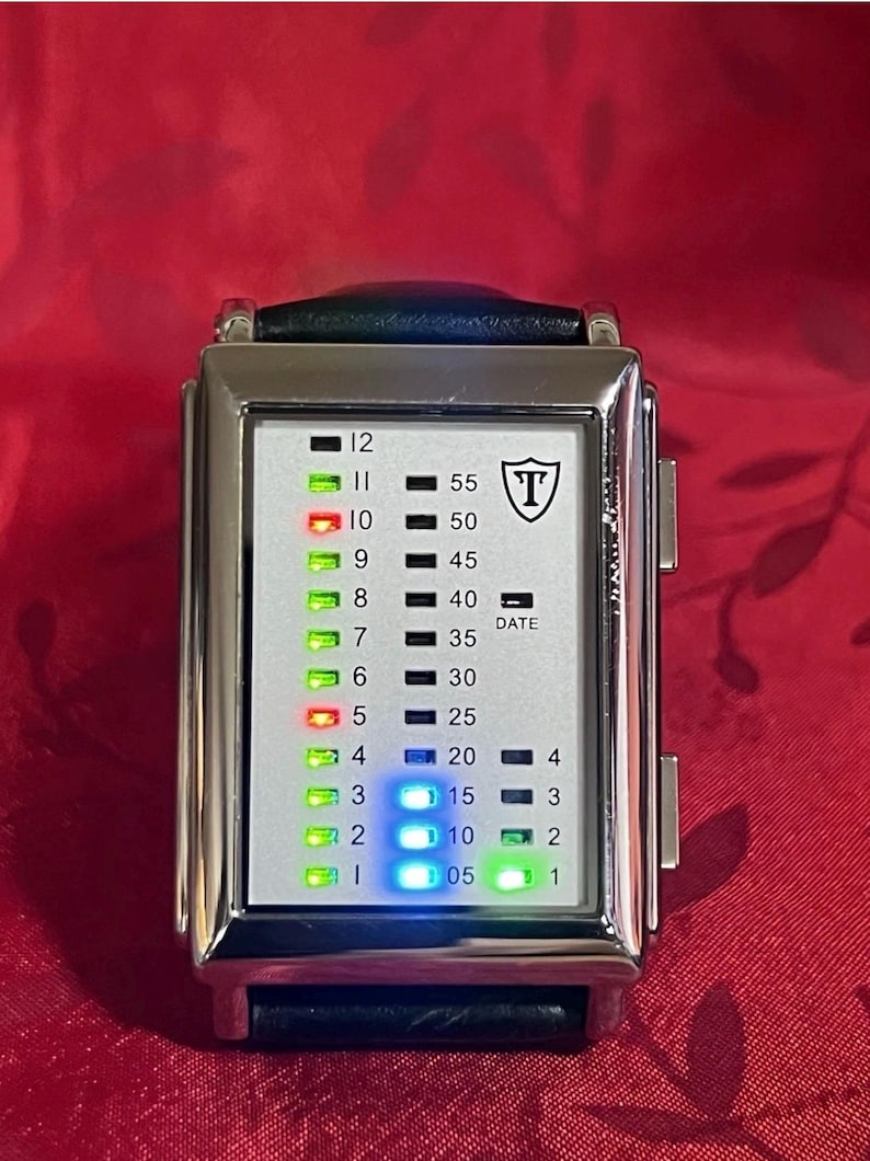 May include: A rectangular digital watch with a silver-colored case and a black leather band. The watch face displays time with a series of colored lights and numbers. The watch has the letter "T" inside a shield logo.