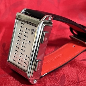 May include: A rectangular, silver-toned digital watch with a black leather strap and red underside. The watch face displays time with illuminated dots and numbers. The watch has "SET" and "MODE" buttons on the side.