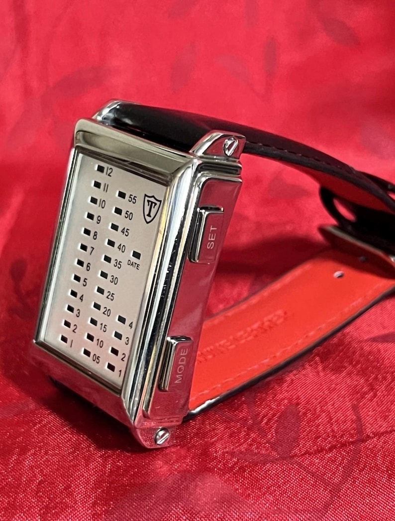 May include: A rectangular, silver-toned digital watch with a black leather strap and red underside. The watch face displays time with illuminated dots and numbers. The watch has "SET" and "MODE" buttons on the side.