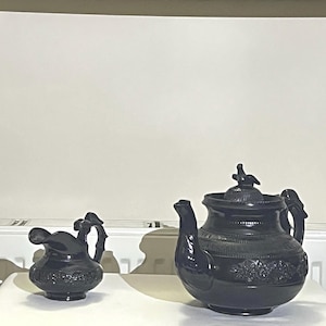 May include: A black ceramic tea set, featuring a teapot with a bird-shaped lid finial and a creamer. The teapot has a curved spout and a decorative floral pattern. The creamer has a curved handle and a similar floral design.