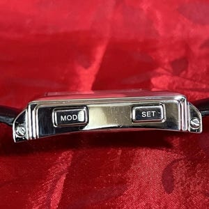 May include: A silver digital watch with a black leather band. The watch face has two buttons labeled "MOD" and "SET". The watch is rectangular with a polished finish. The background is a red fabric.