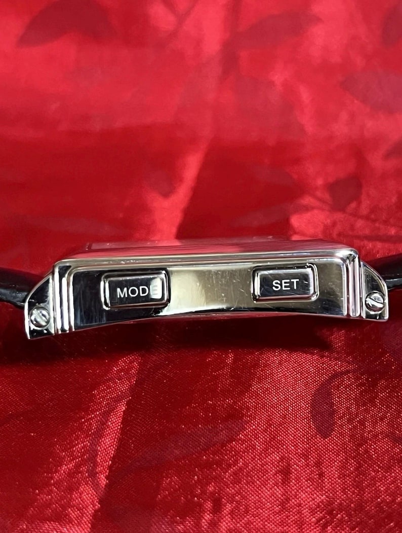 May include: A silver digital watch with a black leather band. The watch face has two buttons labeled "MOD" and "SET". The watch is rectangular with a polished finish. The background is a red fabric.