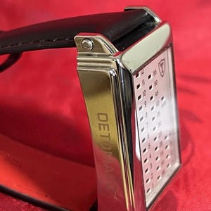 May include: A rectangular wristwatch with a silver-colored case and a black leather strap. The watch face displays a grid of numbers and a logo. The word "DETOMASO" is printed on the side of the watch. The background is red.