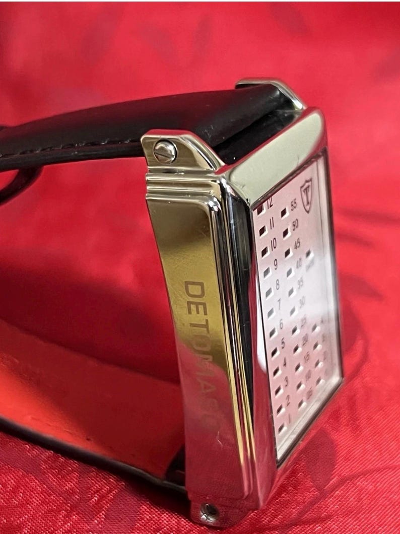 May include: A rectangular wristwatch with a silver-colored case and a black leather strap. The watch face displays a grid of numbers and a logo. The word "DETOMASO" is printed on the side of the watch. The background is red.