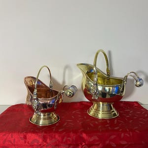 May include: Two antique brass and copper sugar bowls with handles. The bowls have a shiny finish and are set on a red tablecloth. The bowls have a classic design, with a handle and a spout.