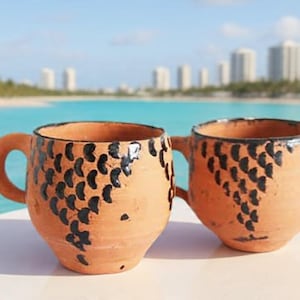 Könnte beinhalten: Zwei handgefertigte Terrakotta-Becher mit schwarzen abstrakten Designs. Die Becher haben ein rustikales, erdiges Aussehen mit einer natürlichen Tonfarbe. Die Becher stehen auf einer weißen Oberfläche mit türkisfarbenem Wasser und einer Skyline im Hintergrund.