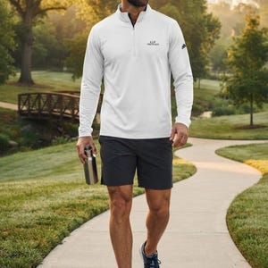 Peut inclure: Un haut à manches longues blanc avec fermeture éclair au col, arborant un petit logo noir sur la poitrine et la manche, associé à un short de sport gris foncé et des chaussures de course grises. Une gourde métallique est tenue à la main.