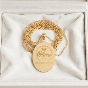 May include: A gold-toned oval pendant necklace featuring an engraved crown, the name Olivia, a small heart, and the text Beauty Has A Name. The pendant hangs from a delicate gold-toned chain, presented inside a white velvet-lined jewellery box.