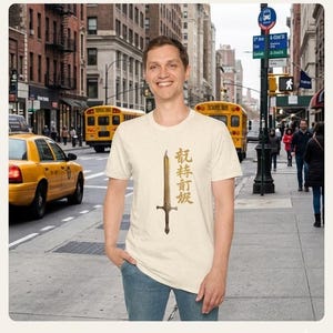 Peut inclure: T-shirt crème avec un motif d'épée dorée et des caractères asiatiques. La personne porte le t-shirt dans une rue de la ville avec des bus scolaires jaunes et des taxis en arrière-plan.