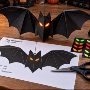 May include: A black paper bat craft kit featuring a 3D assembled bat with glowing orange eyes, a printed Bat Template sheet with cut and fold lines, a pair of black scissors, and a sheet of adhesive eye stickers in orange, red, and green.