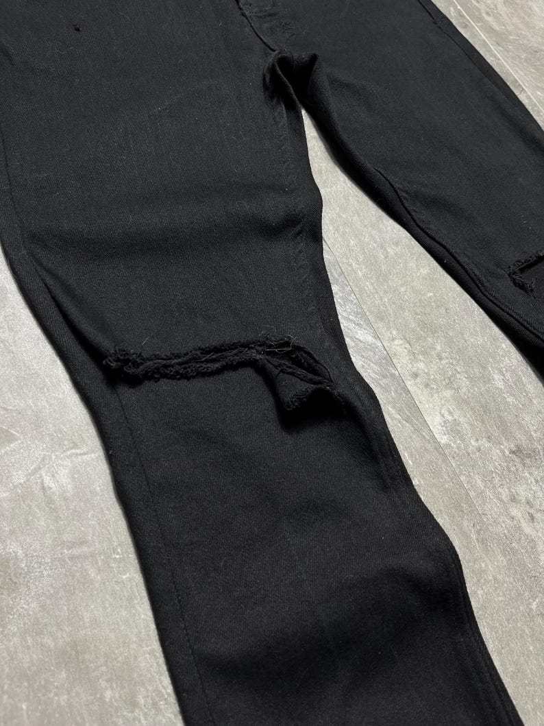 Op de afbeelding: Close-up van een zwarte denim jeans met distressed details. De jeans heeft gescheurde secties bij de knie&euml;n. De stof lijkt een medium-gewicht denim te zijn. Klassiek vijf-zakken ontwerp.