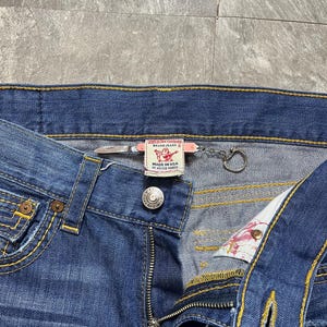 May include: Close-up of blue denim jeans with yellow stitching. The jeans have a button closure, a zipper, and a small metal ring attached to the waistband. A label reads "MADE IN USA OF MEXICO FABRIC".