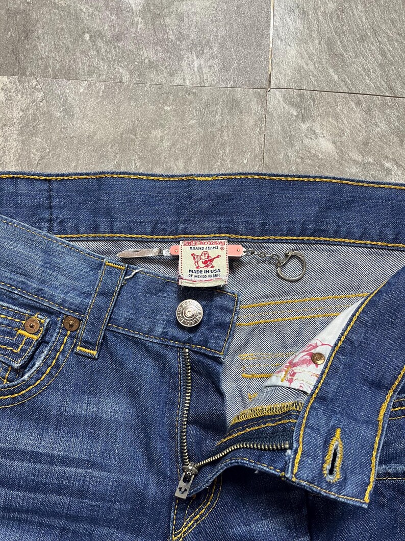 May include: Close-up of blue denim jeans with yellow stitching. The jeans have a button closure, a zipper, and a small metal ring attached to the waistband. A label reads "MADE IN USA OF MEXICO FABRIC".