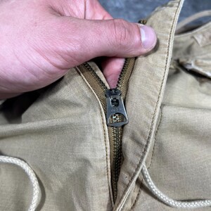 May include: A pair of tan trousers featuring a metal zipper closure with the word IDEAL embossed on the pull tab. The garment is constructed from a woven fabric and includes a light-colored drawstring cord integrated into the waistband area.