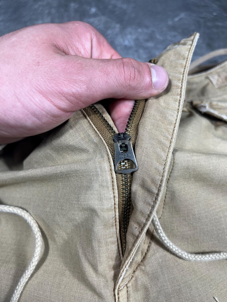 May include: A pair of tan trousers featuring a metal zipper closure with the word IDEAL embossed on the pull tab. The garment is constructed from a woven fabric and includes a light-colored drawstring cord integrated into the waistband area.