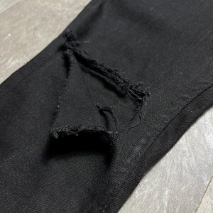 Op de afbeelding: Close-up van een zwarte denim jeans met een gescheurde knie. De stof is donker en getextureerd, met een zichtbare scheur die de draden blootlegt. De jeans lijkt gedragen te zijn en heeft een casual stijl.