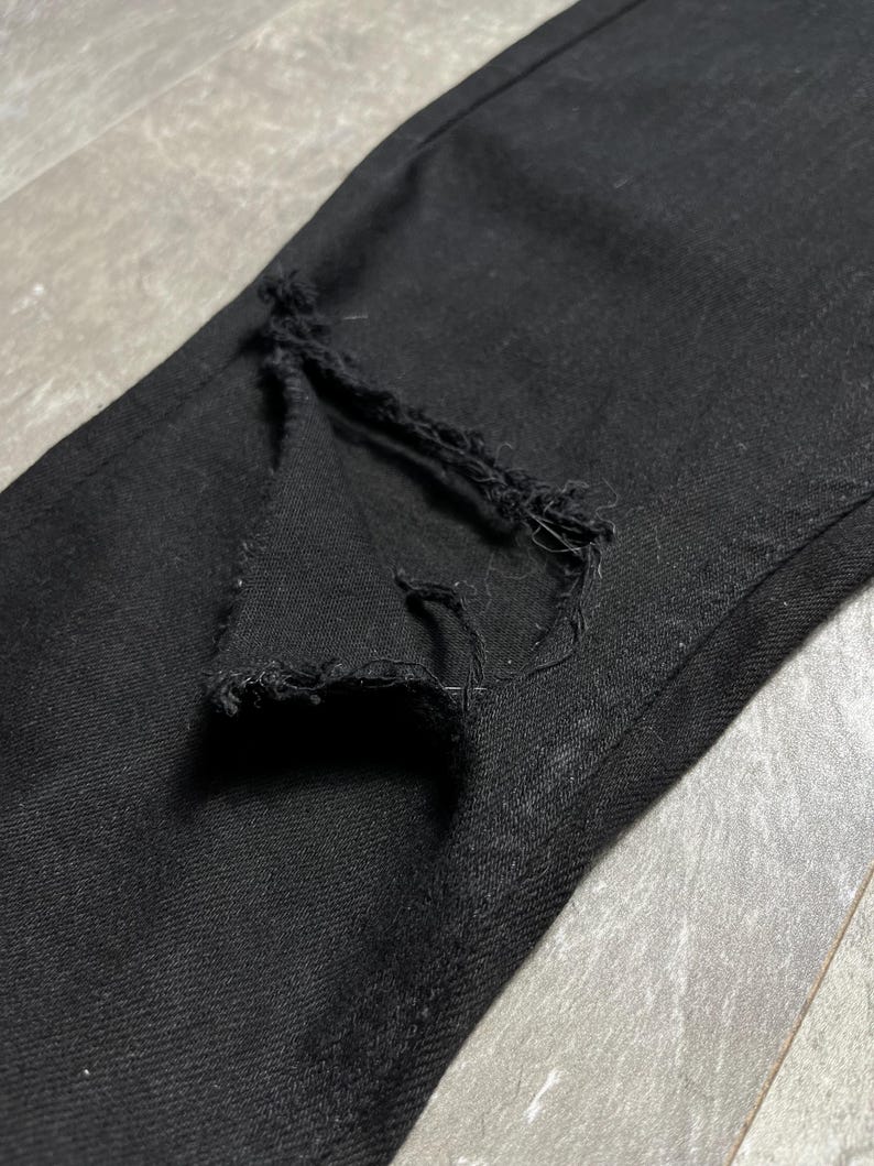 Op de afbeelding: Close-up van een zwarte denim jeans met een gescheurde knie. De stof is donker en getextureerd, met een zichtbare scheur die de draden blootlegt. De jeans lijkt gedragen te zijn en heeft een casual stijl.