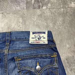 May include: Close-up of blue denim jeans with yellow stitching. The back pocket features a silver button and a decorative stitch design. A rectangular label reads "TRUE RELIGION" with additional text.