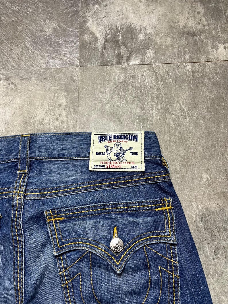 May include: Close-up of blue denim jeans with yellow stitching. The back pocket features a silver button and a decorative stitch design. A rectangular label reads "TRUE RELIGION" with additional text.