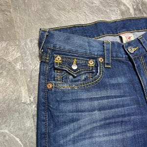 May include: Close-up of blue denim jeans with yellow stitching. The jeans feature a small front pocket with a button closure and a metal rivet. The brand tag is visible on the back pocket.