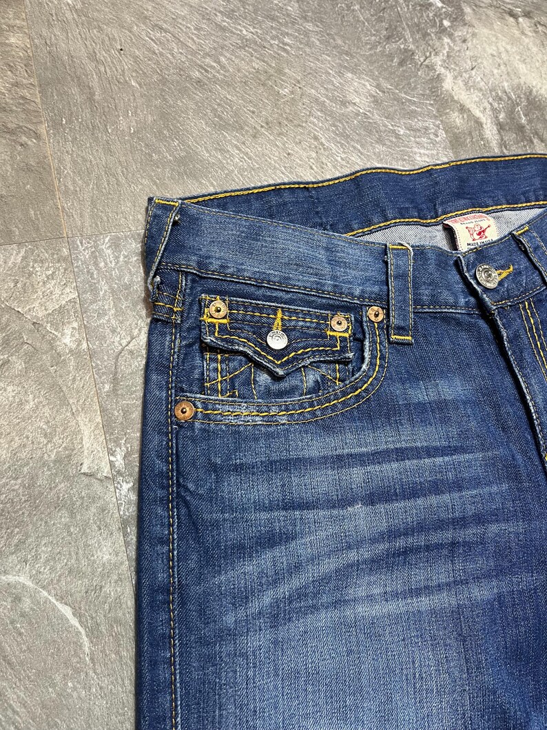 May include: Close-up of blue denim jeans with yellow stitching. The jeans feature a small front pocket with a button closure and a metal rivet. The brand tag is visible on the back pocket.
