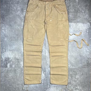 May include: Tan cargo pants featuring a drawstring waist, two slanted front pockets, two large side cargo pockets with flaps, and reinforced knee panels. The leg openings include adjustable drawstrings, and additional fabric ties extend from the side.