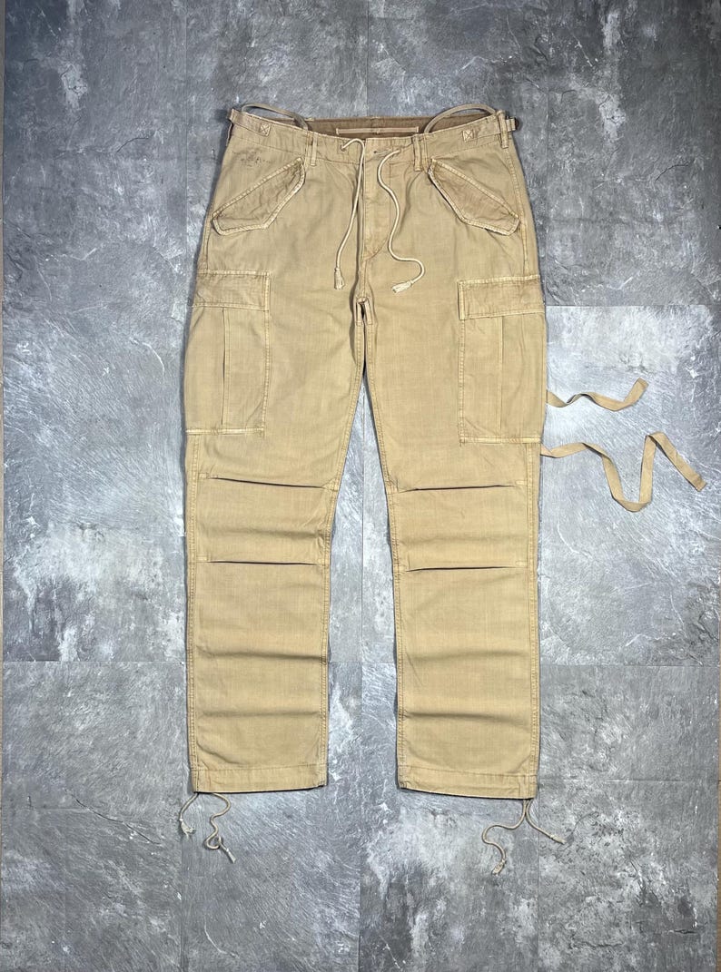 May include: Tan cargo pants featuring a drawstring waist, two slanted front pockets, two large side cargo pockets with flaps, and reinforced knee panels. The leg openings include adjustable drawstrings, and additional fabric ties extend from the side.