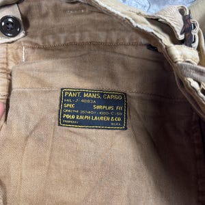 May include: Tan cargo pants featuring a black rectangular interior label with yellow text reading PANT. MANS. CARGO, MIL-J-4883A, SPEC SURPLUS FIT, QMIC THI 35740.1-1020-C-59, POLO RALPH LAUREN & CO. PROPERTY U.S.R.L. The pants have a button closure.