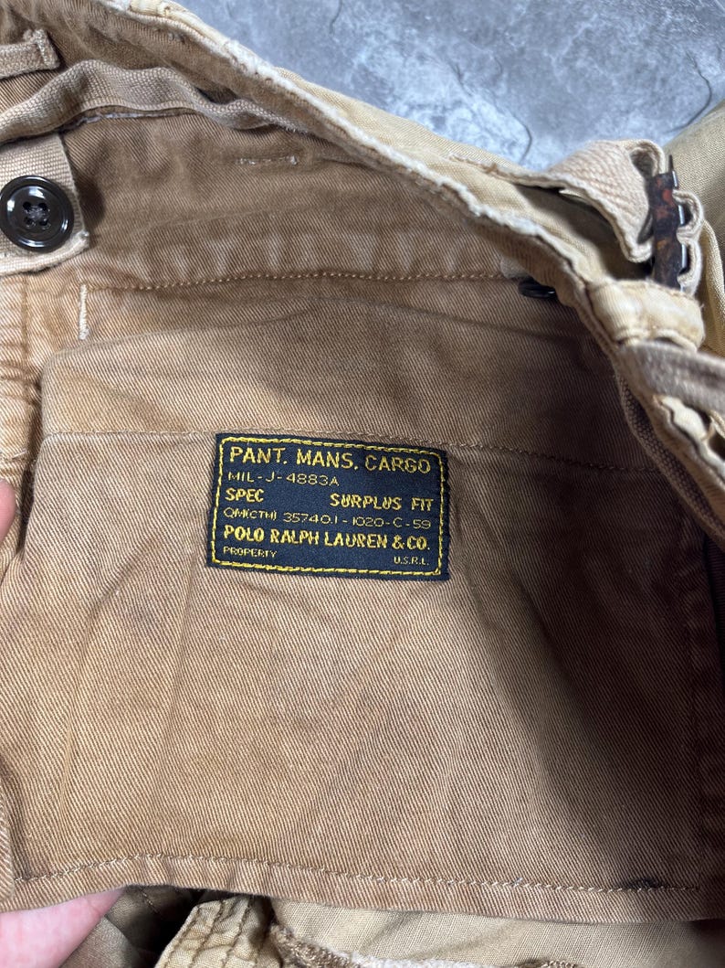 May include: Tan cargo pants featuring a black rectangular interior label with yellow text reading PANT. MANS. CARGO, MIL-J-4883A, SPEC SURPLUS FIT, QMIC THI 35740.1-1020-C-59, POLO RALPH LAUREN & CO. PROPERTY U.S.R.L. The pants have a button closure.