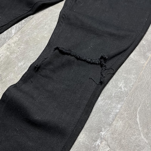 Op de afbeelding: Close-up van een zwarte spijkerbroek met een beschadigde knie. De jeans is effen zwart van kleur en lijkt gemaakt te zijn van stevig denim materiaal. De knie heeft een grote scheur met gerafelde randen.