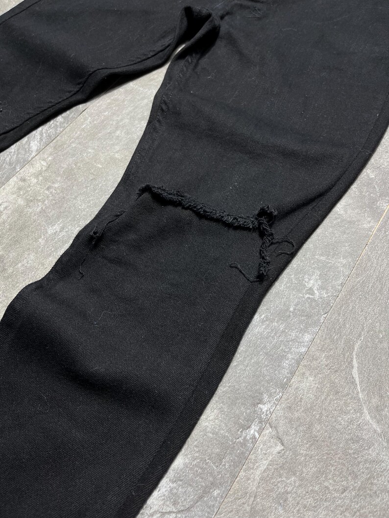 Op de afbeelding: Close-up van een zwarte spijkerbroek met een beschadigde knie. De jeans is effen zwart van kleur en lijkt gemaakt te zijn van stevig denim materiaal. De knie heeft een grote scheur met gerafelde randen.