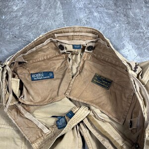 May include: Tan cargo pants featuring a Polo by Ralph Lauren label on the interior pocket. The interior includes a black military-style spec tag with white text reading PANT, MANS, CARGO, SURPLUS, and Polo Ralph Lauren & Co. The pants have a zipper.