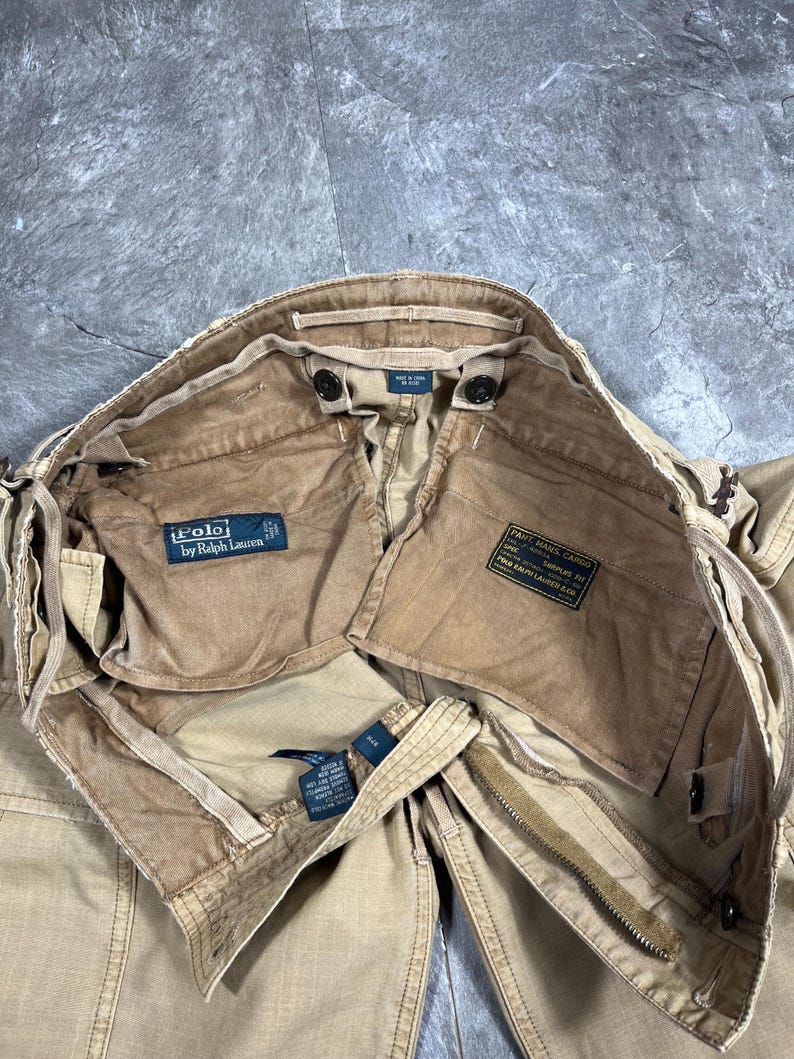 May include: Tan cargo pants featuring a Polo by Ralph Lauren label on the interior pocket. The interior includes a black military-style spec tag with white text reading PANT, MANS, CARGO, SURPLUS, and Polo Ralph Lauren & Co. The pants have a zipper.