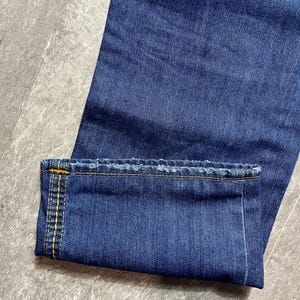 May include: Close-up of blue denim jeans with a folded cuff. The cuff is stitched with yellow thread. The jeans are a medium wash and appear to be new. The fabric has a textured appearance.