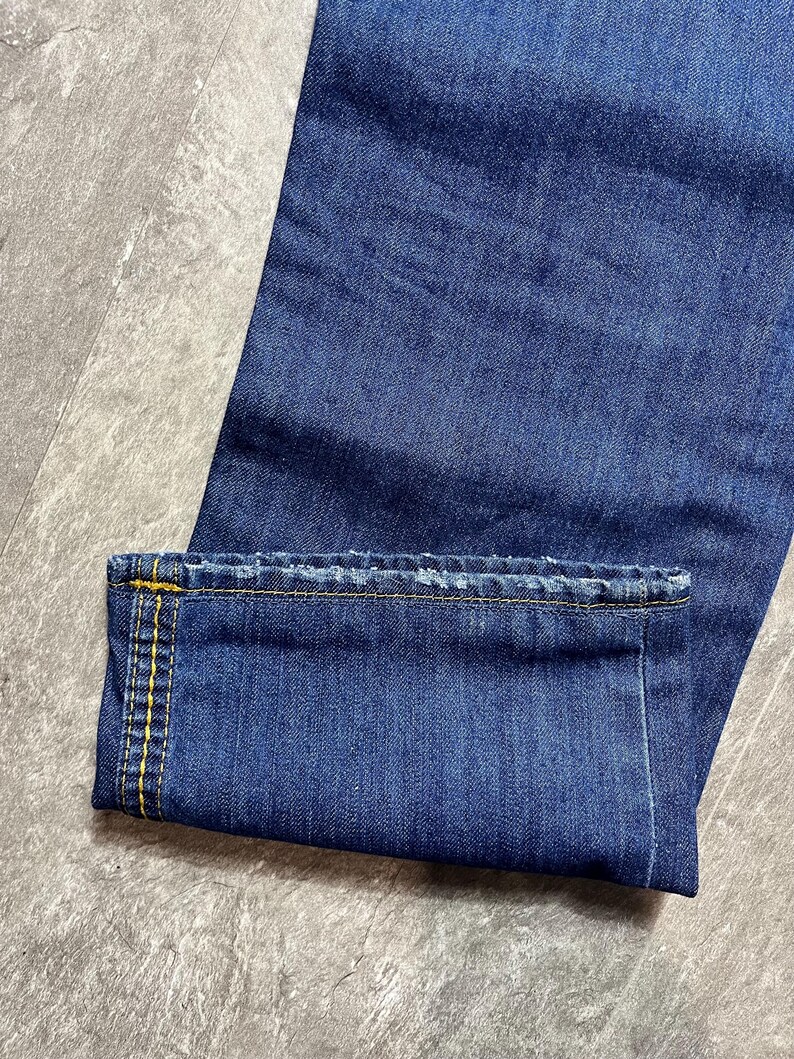 May include: Close-up of blue denim jeans with a folded cuff. The cuff is stitched with yellow thread. The jeans are a medium wash and appear to be new. The fabric has a textured appearance.