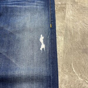 May include: Close-up of blue denim jeans with a distressed patch on the leg. The jeans have a classic five-pocket design, with visible stitching and a faded wash. The denim is a medium blue color.