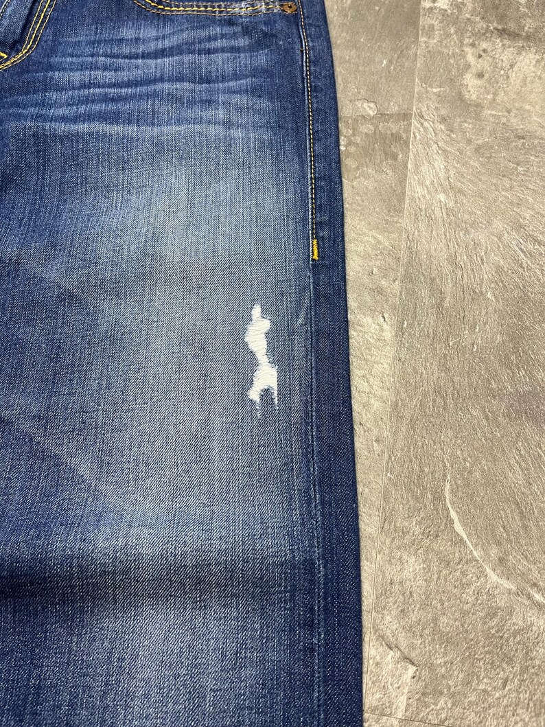 May include: Close-up of blue denim jeans with a distressed patch on the leg. The jeans have a classic five-pocket design, with visible stitching and a faded wash. The denim is a medium blue color.