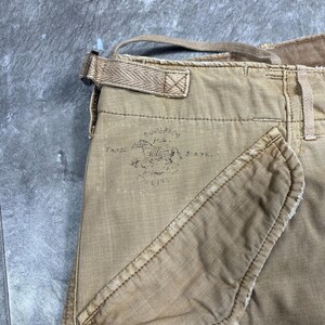 May include: Tan cargo pants featuring a side pocket and a waist adjustment strap with a herringbone pattern. A faded black stamp on the fabric displays a horse graphic with the text Superior Trade Mark, showing visible wear and frayed edges.