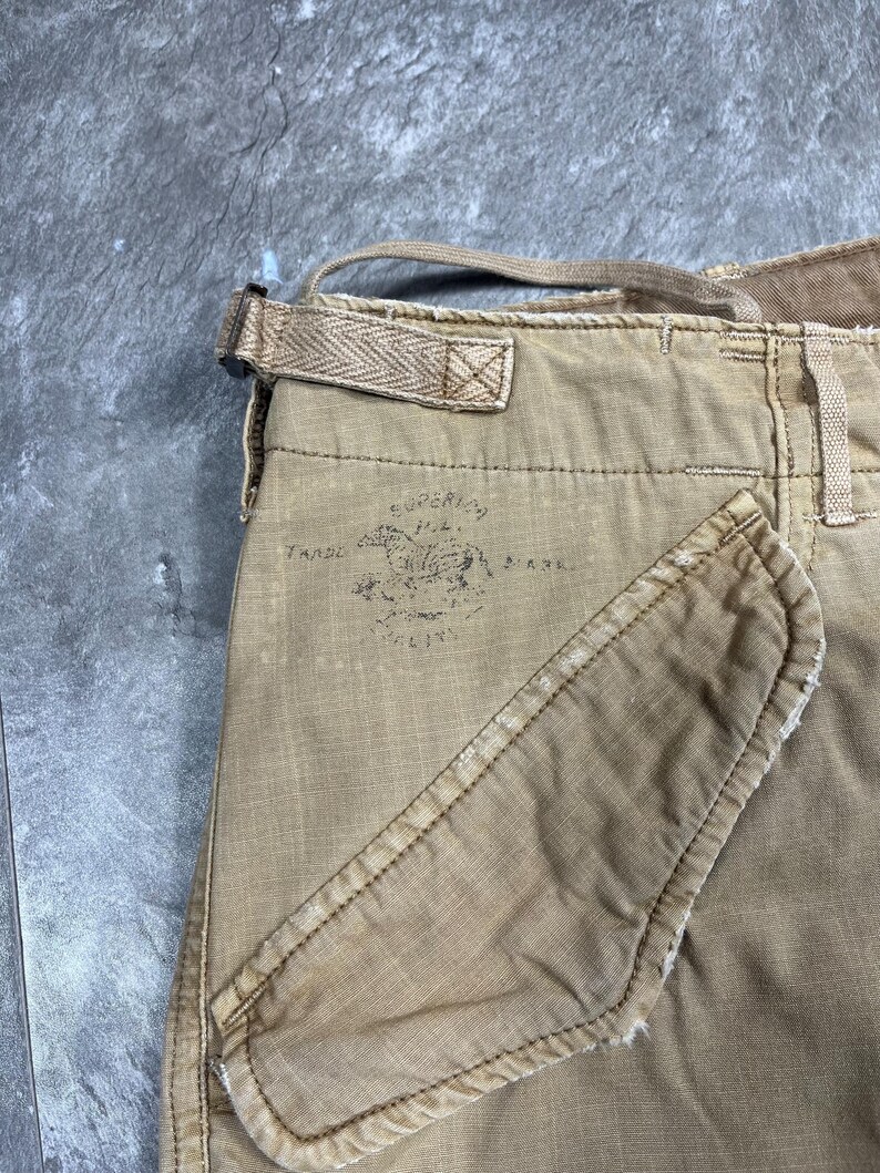 May include: Tan cargo pants featuring a side pocket and a waist adjustment strap with a herringbone pattern. A faded black stamp on the fabric displays a horse graphic with the text Superior Trade Mark, showing visible wear and frayed edges.