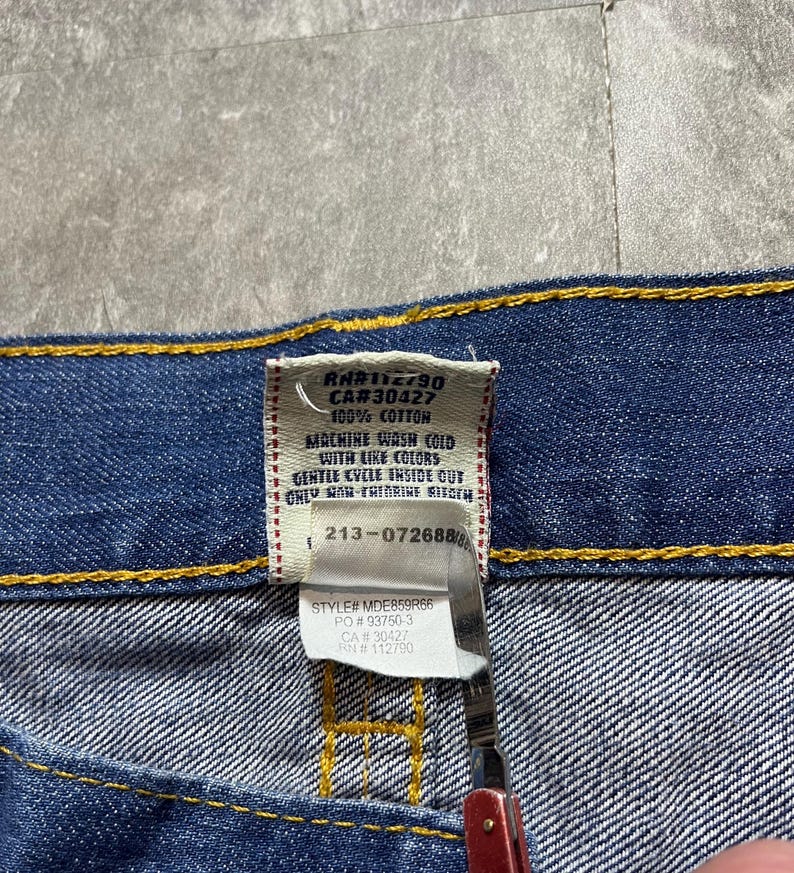 May include: Close-up of a blue denim garment with a tag. The tag displays care instructions and fabric content: 100% cotton. The tag also includes the style number MDE859R66 and RN#112790.