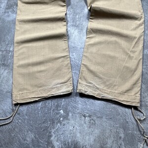 May include: A pair of tan cargo pants featuring wide legs and a straight hem. The pants include side cargo pockets with flap closures and adjustable drawstring ties at the bottom of each leg opening, allowing for a customizable fit at the ankles.