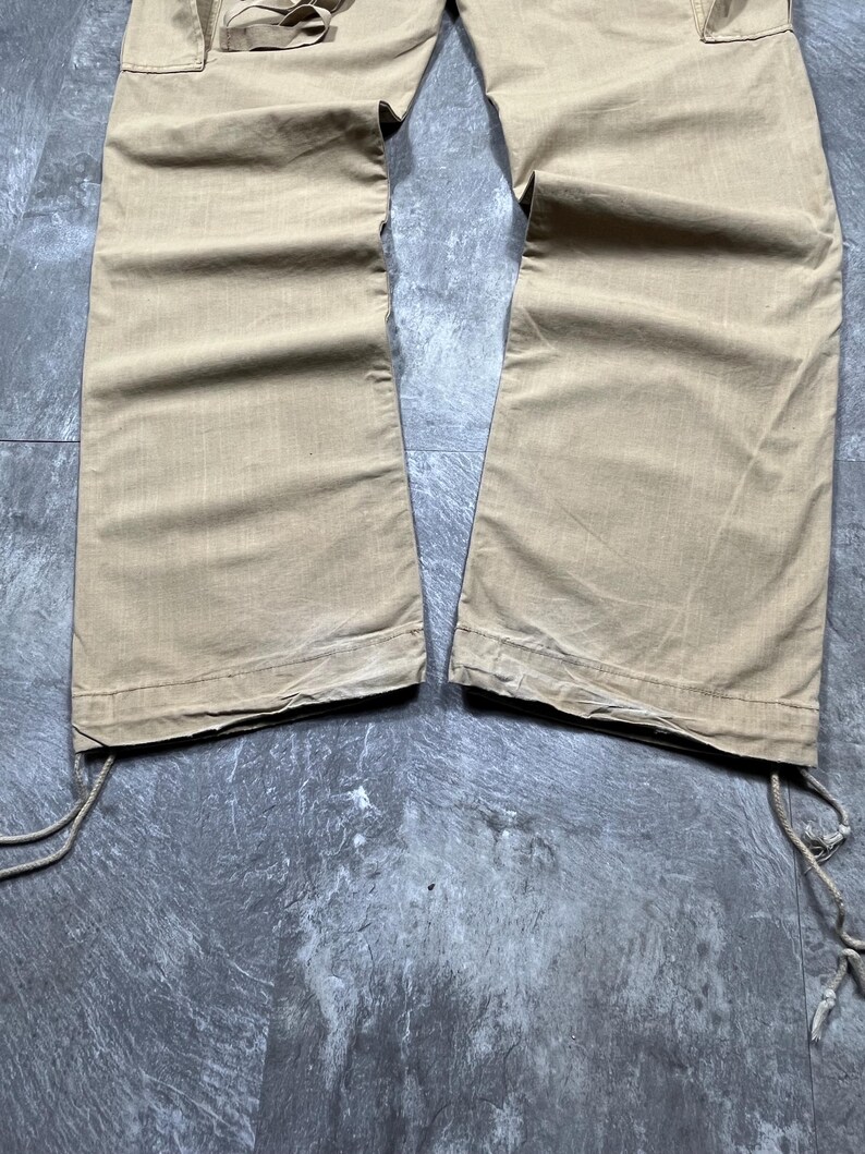May include: A pair of tan cargo pants featuring wide legs and a straight hem. The pants include side cargo pockets with flap closures and adjustable drawstring ties at the bottom of each leg opening, allowing for a customizable fit at the ankles.