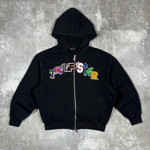 Pode incluir: Casaco preto com zíper, capuz e bolsos. A frente apresenta a palavra "Trapstar" em letras bordadas coloridas. O casaco tem um zíper prateado e um estilo casual.