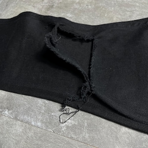 Op de afbeelding: Close-up van een zwarte denim jeans met een distressed, gescheurd detail bij de knie. De stof vertoont tekenen van slijtage, met gerafelde randen en een zichtbaar gat. De jeans heeft een effen zwarte kleur.