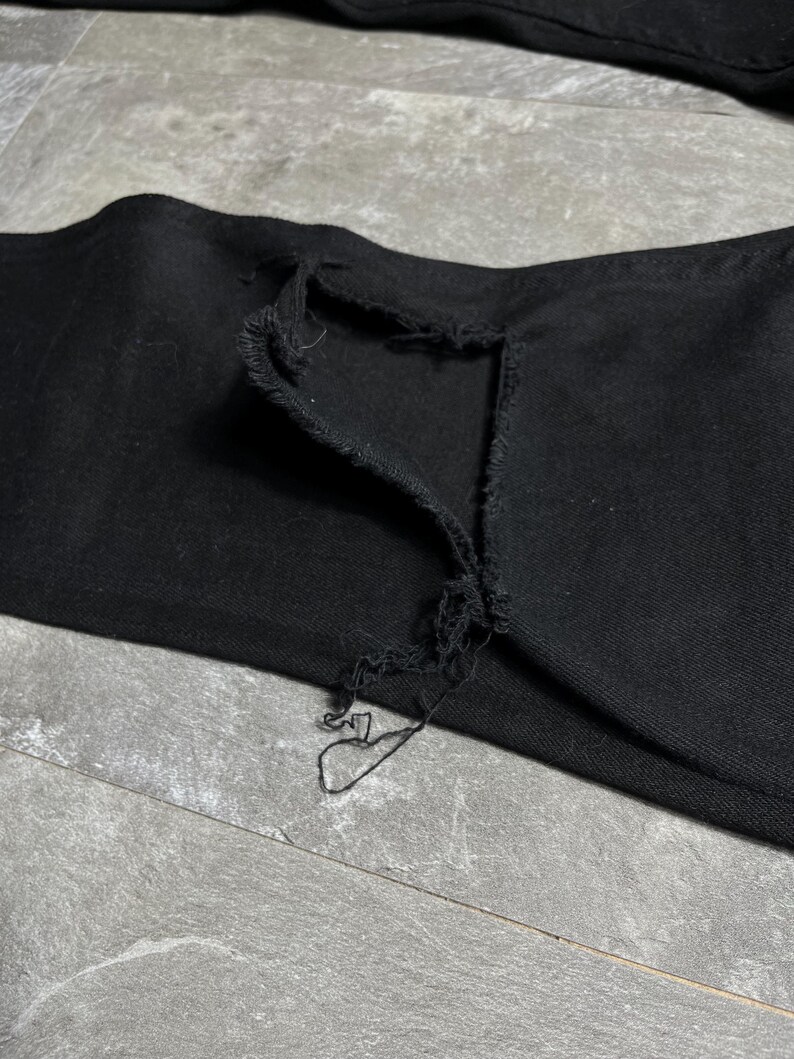 Op de afbeelding: Close-up van een zwarte denim jeans met een distressed, gescheurd detail bij de knie. De stof vertoont tekenen van slijtage, met gerafelde randen en een zichtbaar gat. De jeans heeft een effen zwarte kleur.