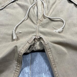 May include: Tan cargo pants featuring a drawstring waist with white cord ties, reinforced stitching at the crotch, and side pockets. The fabric has a visible grid-like texture, and the waistband includes belt loops and a button closure.
