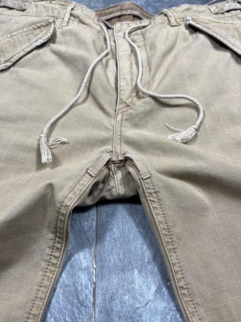 May include: Tan cargo pants featuring a drawstring waist with white cord ties, reinforced stitching at the crotch, and side pockets. The fabric has a visible grid-like texture, and the waistband includes belt loops and a button closure.