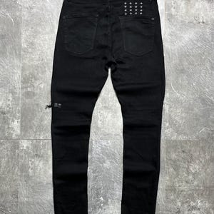 Op de afbeelding: Zwarte denim jeans met een klassiek ontwerp met vijf zakken. De achterzakken hebben een patroon van kleine, witte kruisjes. De jeans heeft een slanke pasvorm en een distressed detail bij de enkel. De jeans worden gepresenteerd op een grijze ondergrond.