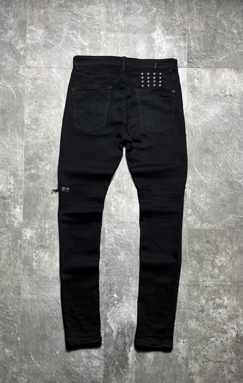 Op de afbeelding: Zwarte denim jeans met een klassiek ontwerp met vijf zakken. De achterzakken hebben een patroon van kleine, witte kruisjes. De jeans heeft een slanke pasvorm en een distressed detail bij de enkel. De jeans worden gepresenteerd op een grijze ondergrond.