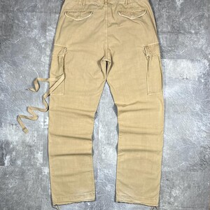 May include: Tan cargo pants featuring two side cargo pockets, two back pockets with flaps, and a waistband with belt loops. The pants include long fabric ties attached to the left cargo pocket and adjustable drawstrings at the bottom hems.